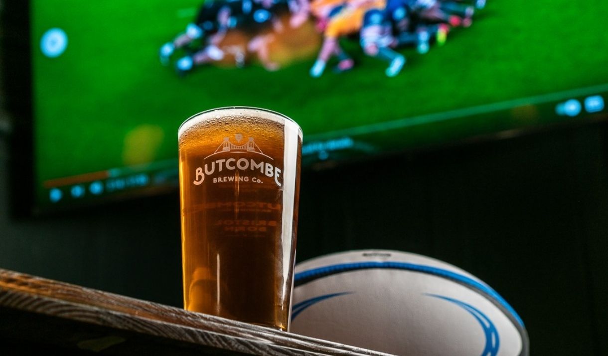 a glass of butcombe beer/cider on a table, next to a rugby ball and a screen playing a rugy match